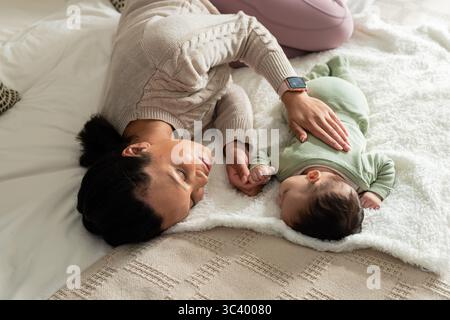 Mother and baby lying on bed in bedroom holding hands on plush blankets with smartwatch detail Stock Photo
