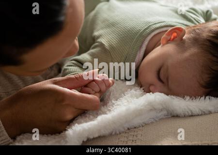Mother and infant daughter lying on bed holding hands with white plush blanket and beige bedspread Stock Photo