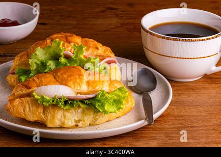 Croissant ham cheese with green vegetable and slice onion in brown spotted ceramic plate with a cup of black coffee and ketchub in a white ceramic bow Stock Photo
