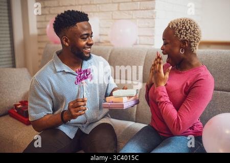 Happy young couple celebrating woman's birthday at home. Boyfriend is giving present to his girlfriend. Stock Photo