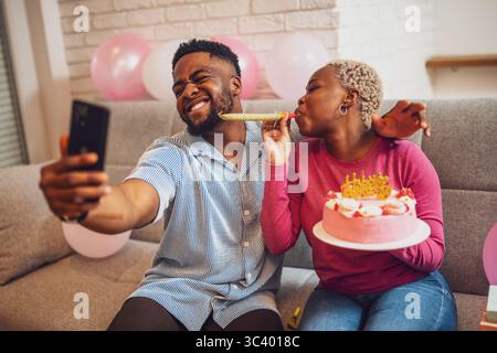 Happy young couple celebrating woman's birthday at home. Woman is holding cake and they are having fun with birthday trumpet while taking selfie. Stock Photo