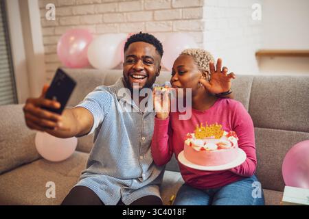 Happy young couple celebrating woman's birthday at home. Woman is holding cake and they are having fun with birthday trumpet while taking selfie. Stock Photo