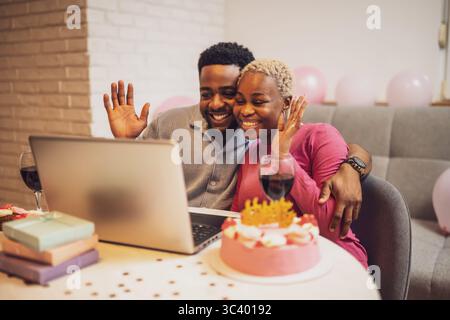Happy young couple celebrating woman's birthday at home. They are having video call with family. Stock Photo
