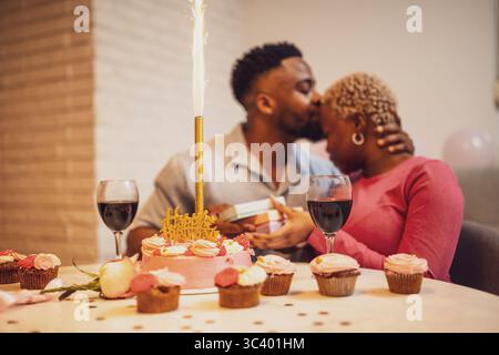 Happy young couple celebrating woman's birthday at home. They are embracing and boyfriend is giving present to his girlfriend. Stock Photo