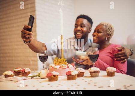 Happy young couple celebrating woman's birthday at home. They are embracing and taking selfie. Stock Photo