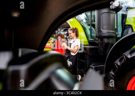 Female mechanic using mobile phone next to tractor in farm equipment yard Stock Photo