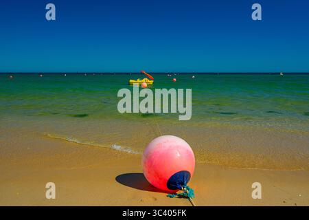 A round red signal buoy on the white sand surface of a beach with crystal clear waters, with a yellow pedal boat in the background on a sunny summer Stock Photo