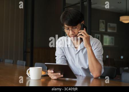 Asian man wearing eyeglasses talking on the phone,  looking data information at tablet, work in the morning on the wooden dining table with hot coffee Stock Photo