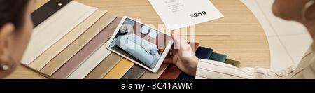 Two young adult Caucasian women sitting at round table reviewing sofa options on digital tablet, comparing fabric swatches and discussing furniture selection in store setting Stock Photo