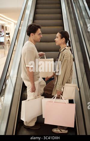 Young adult Caucasian man and young adult Caucasian woman standing on escalator holding shopping bags facing each other in furniture store engaging in conversation Stock Photo