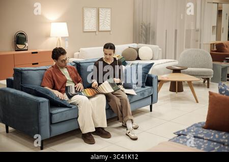 Caucasian man and young adult Caucasian woman sitting on sofa examining fabric swatches together, discussing material options in furniture store showroom setting Stock Photo