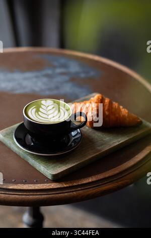 Cup of matcha latte with beautiful latte art served alongside a fresh croissant on a rustic wooden table in a cozy coffee shop. Warm and inviting cafe Stock Photo