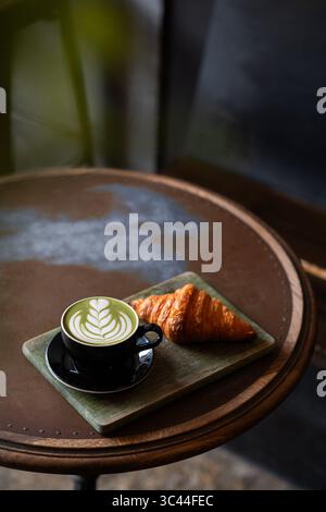 Cup of matcha latte with beautiful latte art served alongside a fresh croissant on a rustic wooden table in a cozy coffee shop. Warm and inviting cafe. Stock Photo