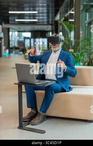 Mature businessman programmer software developer using headset app making a presentation. Watching virtual reality 3D 360 video. Bearded gray-haired guy in suit in VR goggles working at office lobby. Stock Photo