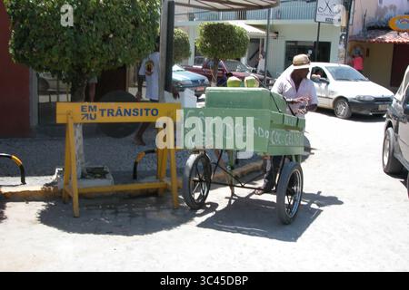 A man wearing a hat is standing next to a cart that says "Coco Gelado" on it. The cart is selling coconuts. There is a yellow sign that says "Em Trans Stock Photo
