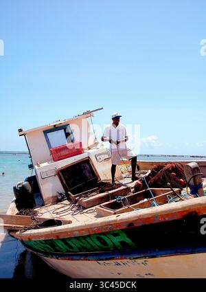 A man stands on a fishing boat, holding a rope. The boat is on the shore, and the ocean is in the background. Stock Photo