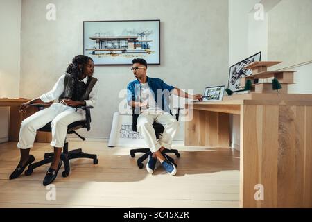 Two young architects discuss project concepts in a stylish design office with creative tools and models visible. Stock Photo