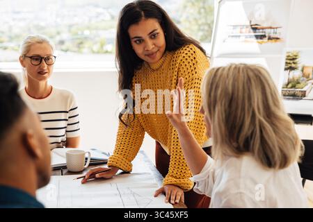 A diverse group of architects, including young women and men, discussing blueprints actively in a modern office setting, showcasing teamwork and profe Stock Photo