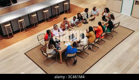 A high-angle view of a diverse group of professionals engaged in a business meeting. The setting is a modern office with a large table and laptops, fo Stock Photo