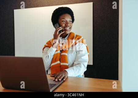 A smiling female entrepreneur is using a smartphone while sitting at a desk with a laptop. She appears confident and engaged in a phone conversation, Stock Photo