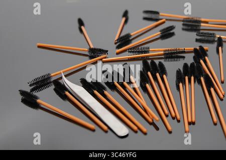 Scattered disposable eyelash brushes and metal tweezers on a glossy black background. Professional tools for lash extension, beauty salon, makeup, and Stock Photo