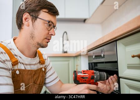 Skilled handyman wearing overalls and safety glasses carefully installs a new oven in a modern kitchen using a cordless drill, ensuring precise and secure placement Stock Photo