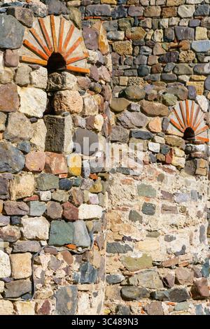 Detailed view of an old stone wall from a medieval fortress with red-brick arched slits and multicolored stones under bright sunlight. structure. Stock Photo