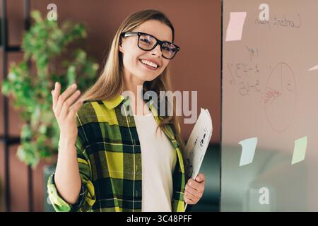 Confident Young Woman Presenting Ideas on a Whiteboard in an Office Setting with a Bright and Engaging Atmosphere Stock Photo