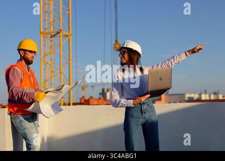 Construction engineers reviewing blueprints and using laptop on site with crane in background, discussing project progress Stock Photo