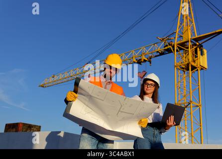 Construction workers reviewing blueprints on a building site with crane in the background, discussing project plans Stock Photo