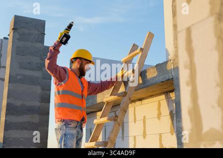 Construction worker using cordless drill on wooden ladder, working on building construction site at sunset Stock Photo