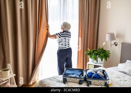 Senior woman closing the curtains on a window in a hotel room Stock Photo