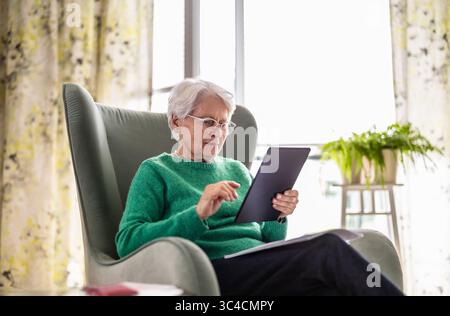 Senior woman using digital tablet while sitting in armchair at home Stock Photo