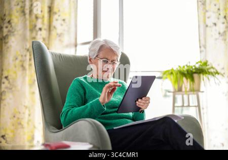 Senior woman using digital tablet while sitting in armchair at home Stock Photo