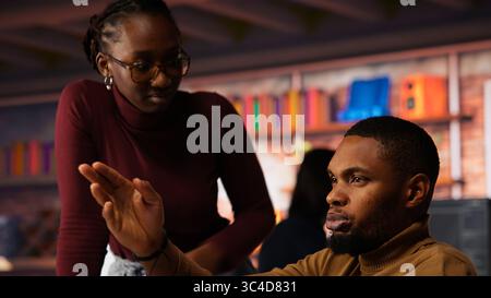 Startup cybersecurity expert in wheelchair looking for company security vulnerabilities on computer, talking with colleague. Man with disability typing on PC and chatting with coworker, camera B Stock Photo