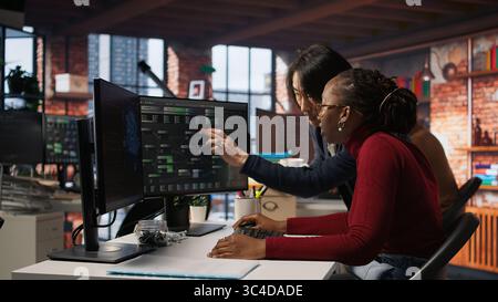 Exhausted AI startup employee rubbing forehead while analyzing datasets on computer, receiving coffee from project CTO manager to get energy and continue coding, camera A Stock Photo