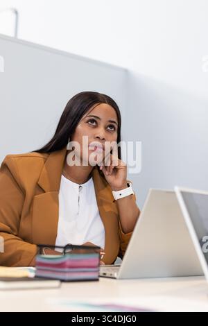 Mid adult African American woman resting chin gazing upward at office with laptop, smartwatch Stock Photo
