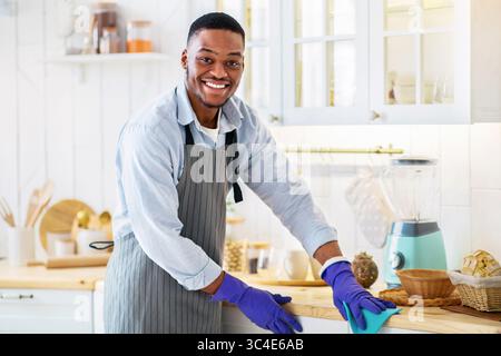 Handsome young black man washing kitchen table with rag at home. Professional sanitary service concept Stock Photo