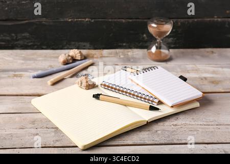 Opened notebooks, pens, markers, hourglass and seashells on wooden table near black wall Stock Photo