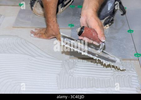 Worker uses trowel to spread adhesive while laying tiles on construction site in broad work day Stock Photo
