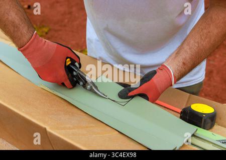 Skilled worker uses scissors to cut green sheet vinyl siding while measuring accurately at wooden surface Stock Photo