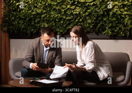 Two business colleagues discuss documents intently on a modern sofa. Professional teamwork, thoughtful expressions. Businessman and assistante . Stock Photo