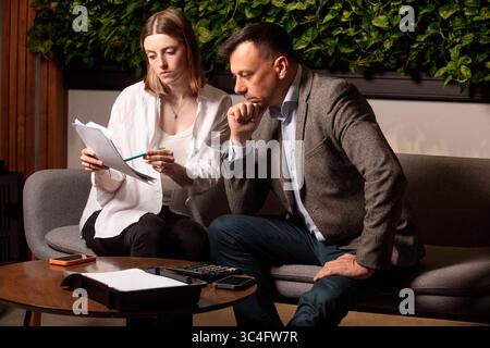 Two business colleagues discuss documents intently on a modern sofa. Professional teamwork, thoughtful expressions. Businessman and assistante . Stock Photo