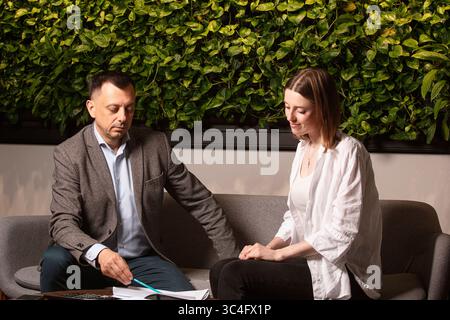 Two business colleagues discuss documents intently on a modern sofa. Professional teamwork, thoughtful expressions. Businessman and assistante . Stock Photo
