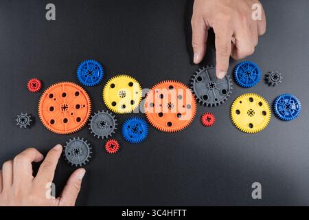 Top view of colorful plastic gears and human hands assembling cogwheels on black background, symbolizing teamwork, engineering, creativity, machine pa Stock Photo