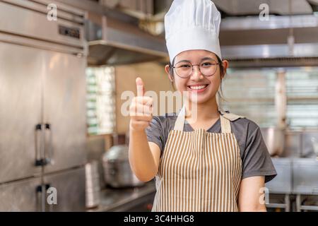 Young female chef smiling and showing a thumbs-up gesture in a professional kitchen environment. She wears a striped apron and a white chef hat, conve Stock Photo