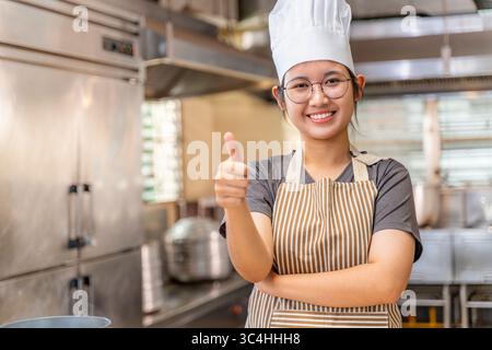 Young female chef smiling and showing a thumbs-up gesture in a professional kitchen environment. She wears a striped apron and a white chef hat, conve Stock Photo