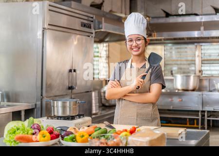 Smiling young female chef in professional kitchen attire, holding a spatula and standing proudly behind a counter filled with fresh vegetables and coo Stock Photo