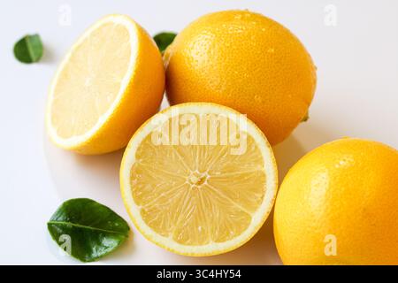 Fresh and vibrant lemons, with one perfectly sliced to reveal its juicy interior, scattered with green leaves on a clean white background. Stock Photo