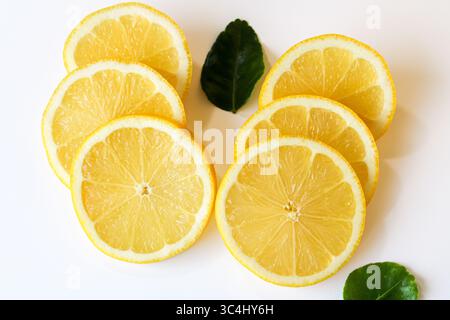 Bright juicy slices of fresh lemon, artfully arranged on a clean white background with scattered green leaves, ready for drinks or garnish. Stock Photo